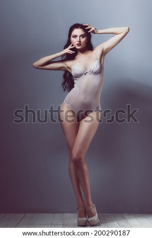 sexy brunette girl in a transparent body beige heels with a beautiful body and long legs and hair developing on a wind in studio on white wooden floor and gray walls