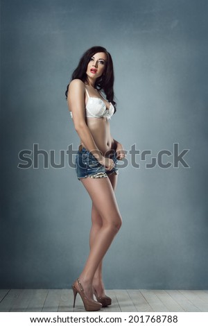 sexy brunette girl with makeup and red lips in shorts and stretches them all white lingerie on her white bra with long legs heels in the studio