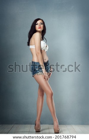 sexy brunette girl with makeup and red lips in shorts and stretches them all white lingerie on her white bra with long legs heels in the studio