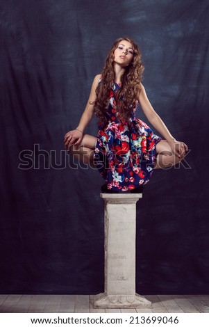 Young beautiful girl sits on a column in the color dress with makeup and long hair in natural black shoes in the studio, sit on the pillar with his legs apart
