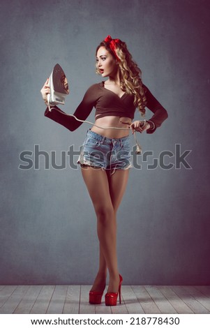 Pin up Young beautiful sexy girl with make-up blonde with a red band in denim shorts housewife with iron in red shoes slender legs