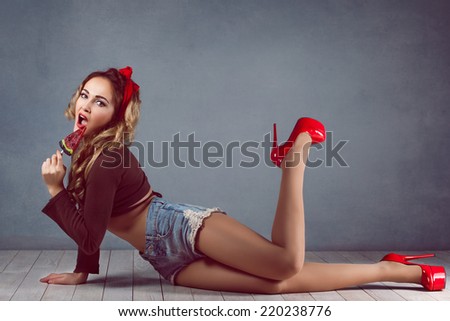 Beautiful young sexy blonde slim figure with a red armband with red lips in denim shorts in red high heels with long legs on the floor sitting watermelon candy pin up emotions