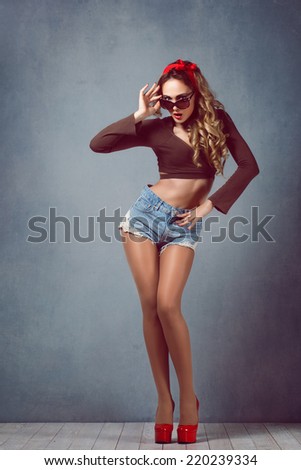 Beautiful young sexy blonde slim figure with a red armband with red lips in denim shorts in red high heels with long legs wearing glasses Pin Up