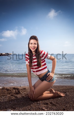 pin up young beautiful girl on the sea in short jeans and striped T-shirt with long hair rocks glasses gulls water clouds, day, wave, dream sand smile with teeth emotions salutes figure