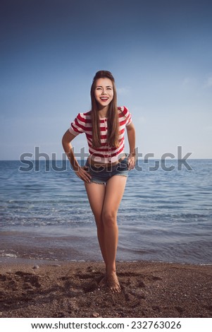 pin up young beautiful girl on the sea in short jeans and striped T-shirt with long hair rocks glasses gulls water clouds, day, wave, dream sand smile with teeth emotions salutes figure