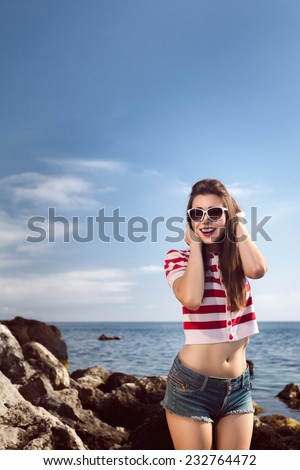 pin up young beautiful girl on the sea in short jeans and striped T-shirt with long hair rocks glasses gulls water clouds, wave, dream figure with glasses emotions, smile with teeth, open mouth