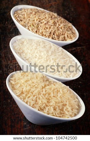Three bowls with different types of rice: brown rice, wild rice and white (jasmine) rice on wood board. Selective focus