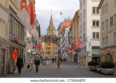 ZURICH, SWITZERLAND - MAY 04, 2013: Cobbled pedestrian street Rennweg with boutique shops, flags on old buildings and St. Peter\'??s church clock tower in Zurich, Switzerland.