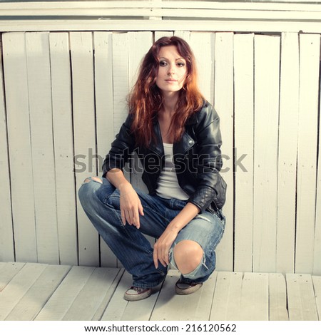 Beautiful young woman in jeans and black leather jacket sitting against grunge wood wall, instagram effect