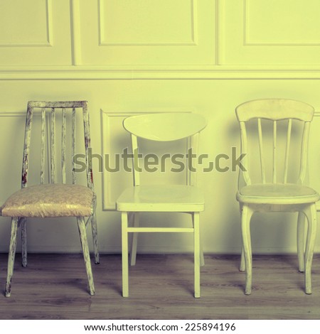 Set of white wooden vintage chairs standing in front of a white wooden wall on light parquet floor. Square toned image, instagram effect