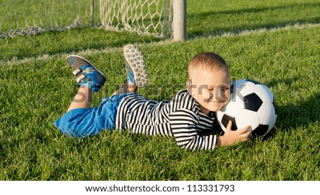 Smiling happy young boy lying on green grass at a sportsfield in evening light with a soccer ball