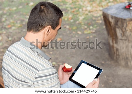 Over the shoulder view of a man sitting on a rural bench using a touchscreen tablet with a blank screen and eating a fresh apple