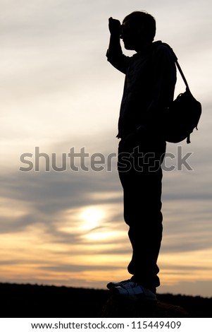 Silhouette of a man with a satchel on his back standing with his hand raised to his forehead looking into distance against a delicate sunset