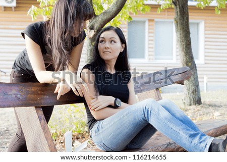 Two young casual woman friends chatting outdoors on a wooden garden bench with one leaning over from behind