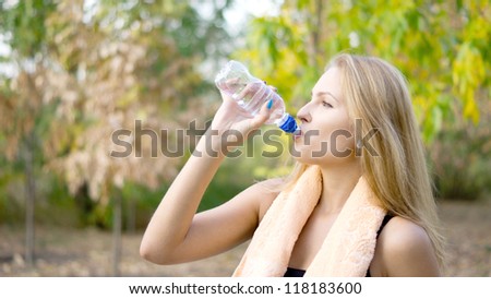 Thirsty woman athlete drinking pure bottled water with a towel around her neck standing in the park