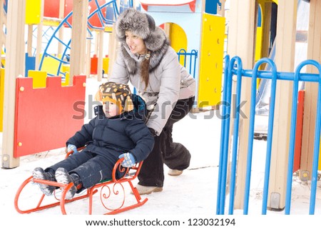 Mother pushing her small son on an ornage sled through the snow on a colourful winter childrens playground
