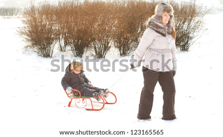 Cute little boy wrapped up warmly against the winter cold riding on a colourful orange sled or toboggan pulled through the snow by his mother