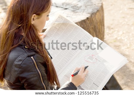 Over the shoulder view of a young female jobseeker reading the classified adverts in a newspaper and ringing them with a felt tip pen