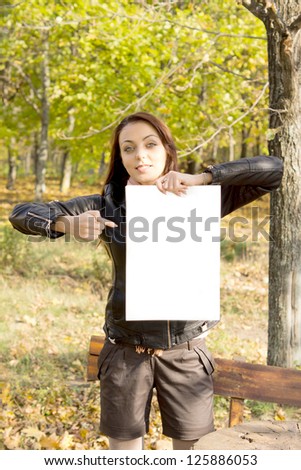 Trendy young woman in shorts holding up a blank sign and pointing to it with her finger while standing alongside a bench in woodland