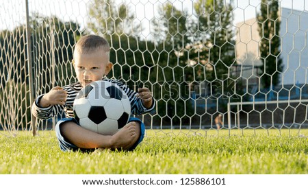Little boy sitting cross legged on the ground in the goalposts with a soccer ball balanced on his lap