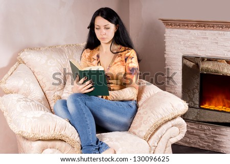 Attractive young woman relaxing in a comfortable armchair reading a book near the blazing fire