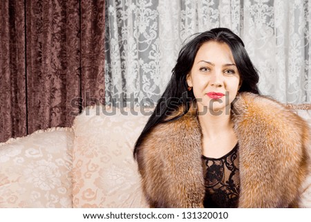 Beautiful young woman with long brunette hair wearing a luxuriant warm fur collar sitting on a sofa in a living room smiling at the camera