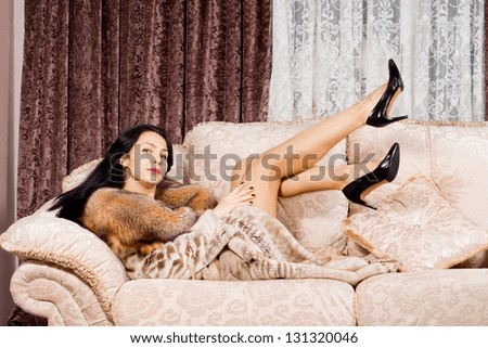 Sexy beautiful woman lying on a cream sofa in a fur coat kicking her long bare legs in the air while looking at the camera