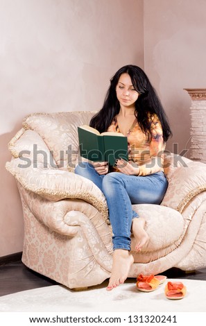 Attractive young brunette woman curled up in a comfortable cream upholstered armchair reading a book with her slip on shoes lying on the carpet beside her