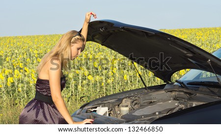 Woman in a smart dress checking under the raised hood of her vehicle as she tries to determine a reason for the breakdown, field of sunflowers in background