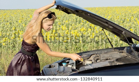 Woman checking the level of the engine oil in her car which has broken down alongside a field of sunflowers