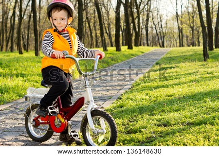Adorable little boy riding his bike along a paved path in a wooded park stopping to stand and pose for the camera in his safety gear consisting of a helmet and orange high visibility jacket