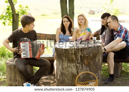 Young man playing music for his friends on an accordion as they enjoy a relaxing day together seated around a rustic tree stump table