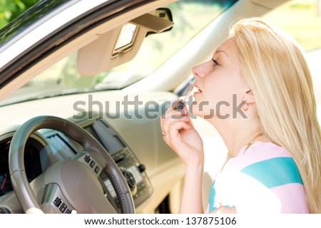 Beautiful young blond woman sitting applying makeup in a car using the mirror in the visor as she freshens up after a journey