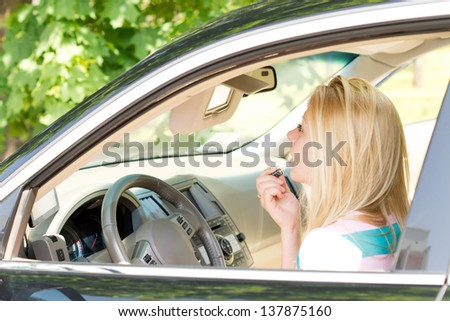 Young attractive blond girl applying lipstick looking in the mirror of the visor in a car.