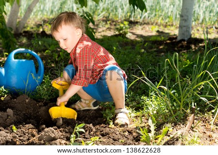 Little boy digging with a colourful yellow toy spade in fresh earth under the shade of a tree in the vegetable garden