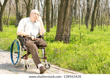 Pensive elderly amputee sitting alone on a rural pathway in his wheelchair with his chin resting on his hand staring into the distance
