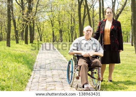 Handicapped elderly man with one leg amputated above the knee sitting in his wheelchair in a wooded park with his wife standing at his side