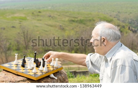 Retired man playing chess in the countryside reaching across to grab a chesspiece to make a move against a scenic valley view