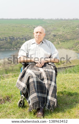 Handicapped man sitting in his wheelchair with a blanket over his legs enjoying the summer sunshine in a quiet rural location with scenic views