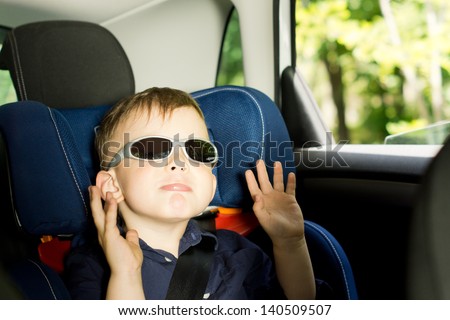 Playful small boy raising arms while sitting in a child car-seat in the back of a car