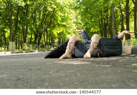 Small boy hidden in a suitcase with his legs protruding over the top as it lies on a rural road under shady trees