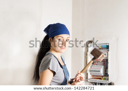 Woman electrician standing in front of an open fuse box with a wooden mallet in one hand and a screwdriver in the other as she contemplates the best solution to the problem