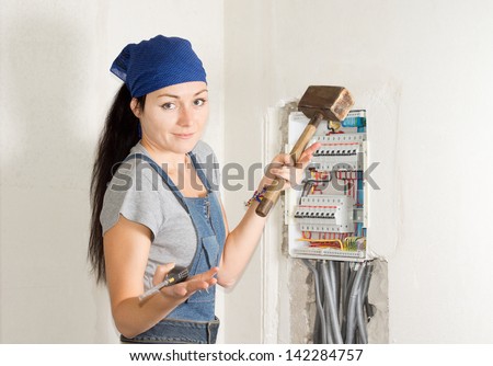 Attractive young female electrician standing in front of an open fuse box with a wooden mallet in one hand and a screwdriver in the other as she contemplates her choices in solving her problem