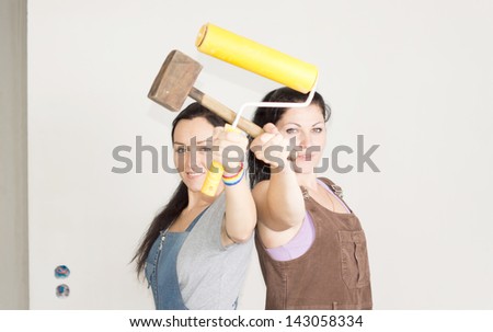 Smiling female friends posing back to back with a wooden mallet and paint roller in their hands as they prepare to start decorating their home