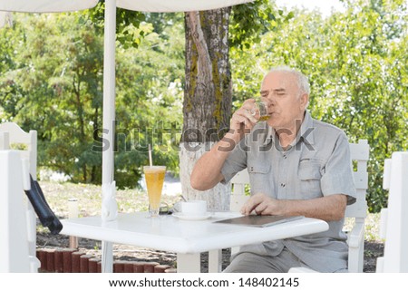 Elderly man sipping a glass of brandy in a snifter as he relaxes on a hot summer day at an outdoor restaurant in the shade of a tree