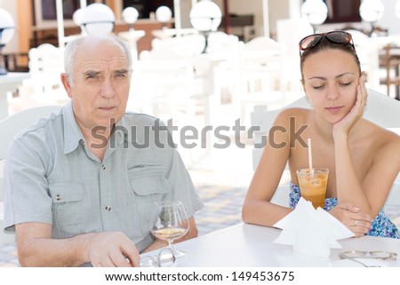 Elderly man and a beautiful younger woman sitting together having drinks at a restaurant at an outdoor table on a hot summer day