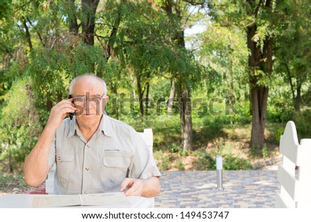 Elderly man chatting on his mobile phone sitting outdoors at a restaurant table in the shade of a tree