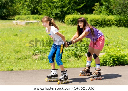 Two attractive teenaged female friends roller skating with the one pulling the other along behind her in a lush green rural park