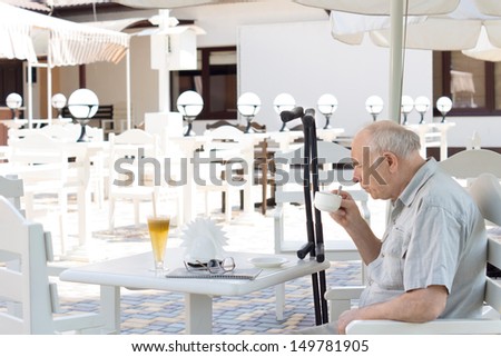 Elderly disabled man enjoying a cup of coffee on his own seated at a table at an outdoor restaurant with his crutches propped alongside