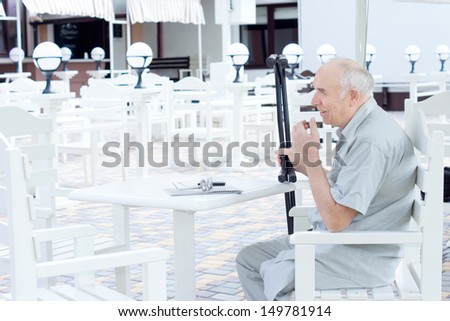 Elderly amputee sitting at a restaurant table outdoors holding his crutches in his hand as he waits for his companion to arrive